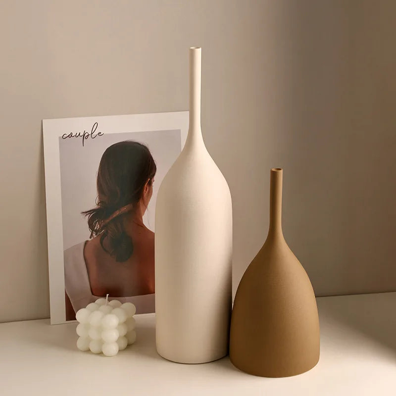 Two ceramic vases, one white and one brown, on a surface with a minimalistic poster in the background.